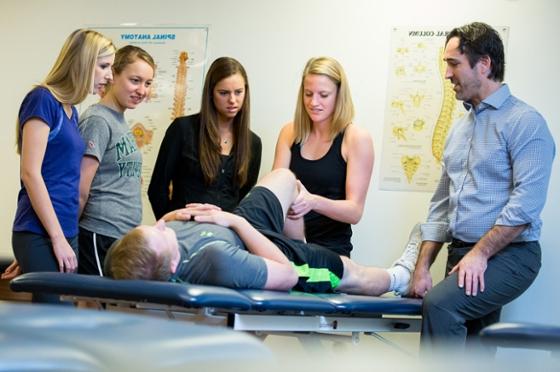 Photo of an instructor and four female physical therapy students at Chatham University surrounding a patient on a table as one student adjusts his leg. 