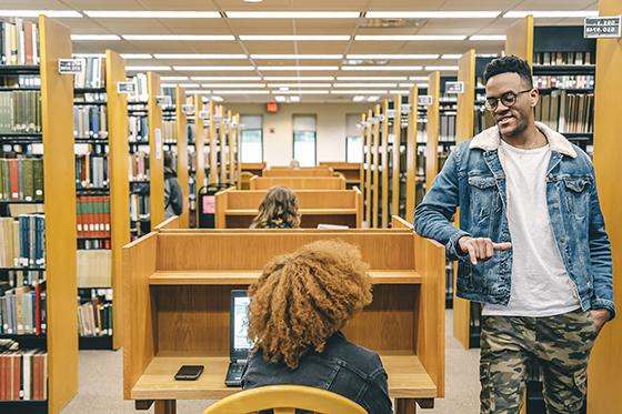 Photo of two students chatting at a cubicle in Jennie Mellon King library, between the stacks of library books.