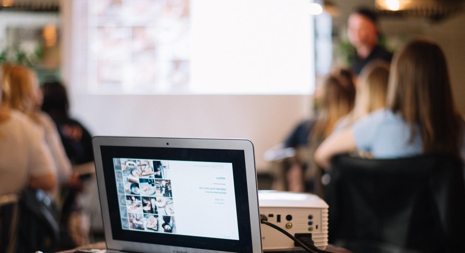 Photo of a projector shining a PowerPoint presentation on screen while connected to a laptop in a classroom. 